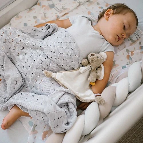 Cream plush dog cuddly toy with dummy holder, held by baby on a bed
