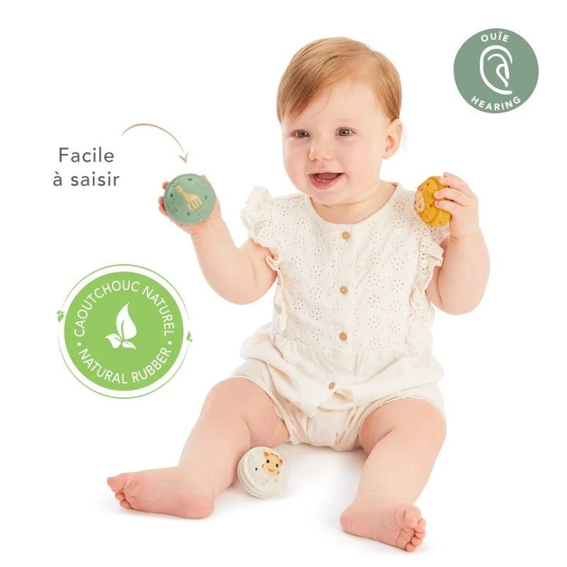 Baby holding yellow and teal musical balls with animal faces while wearing white romper