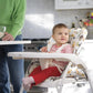 Baby seated in white highchair with patterned fabric and white tray; Joie label on bar.