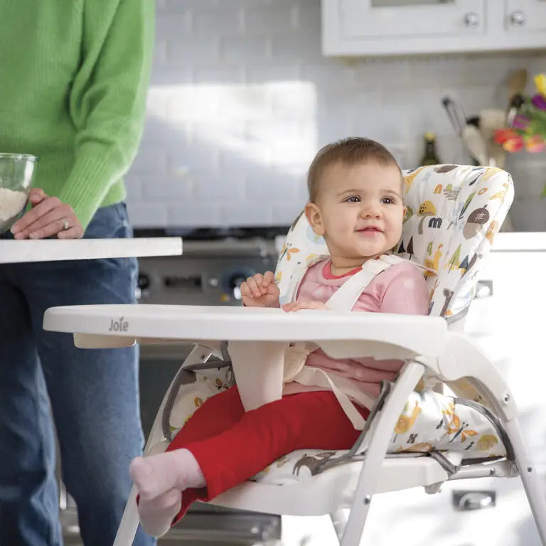 Baby seated in white highchair with patterned fabric and white tray; Joie label on bar.