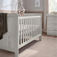 Light gray wood cotbed with vertical rails and two end panels, angled side view