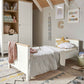 White baby cot with light wood slats and mattress, side view in a nursery