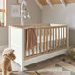 White baby cot with light wood rails, drawer base, and hanging mobile above