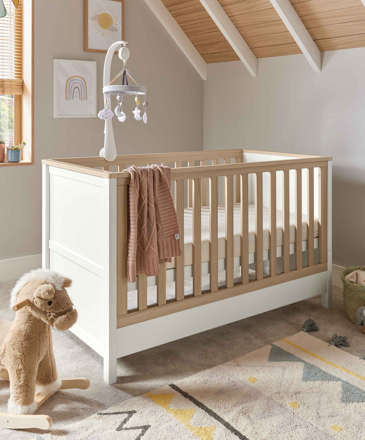 White baby cot with light wood rails, drawer base, and hanging mobile above
