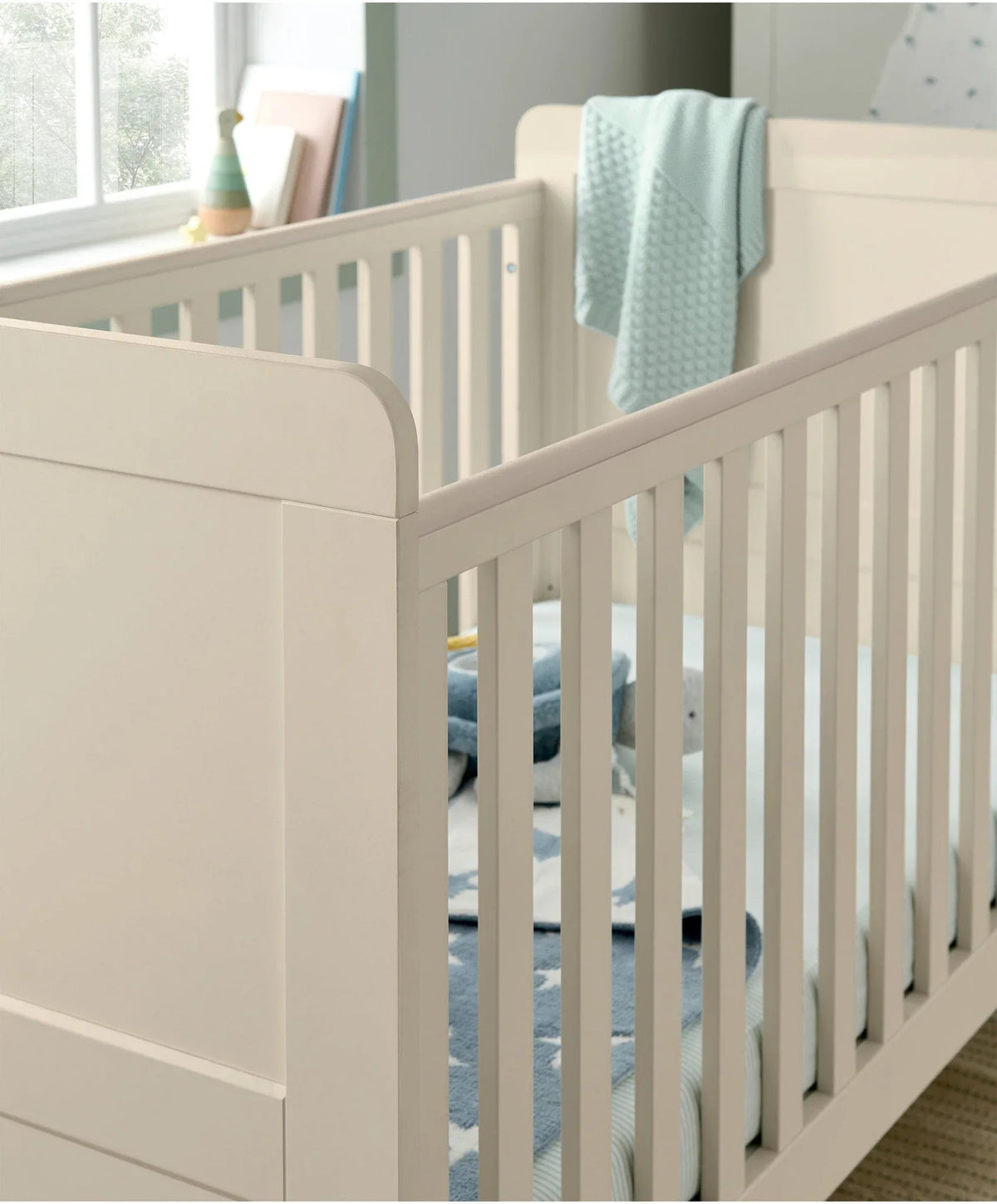 Cream cotbed with vertical slats and raised side rail, with light blue blanket