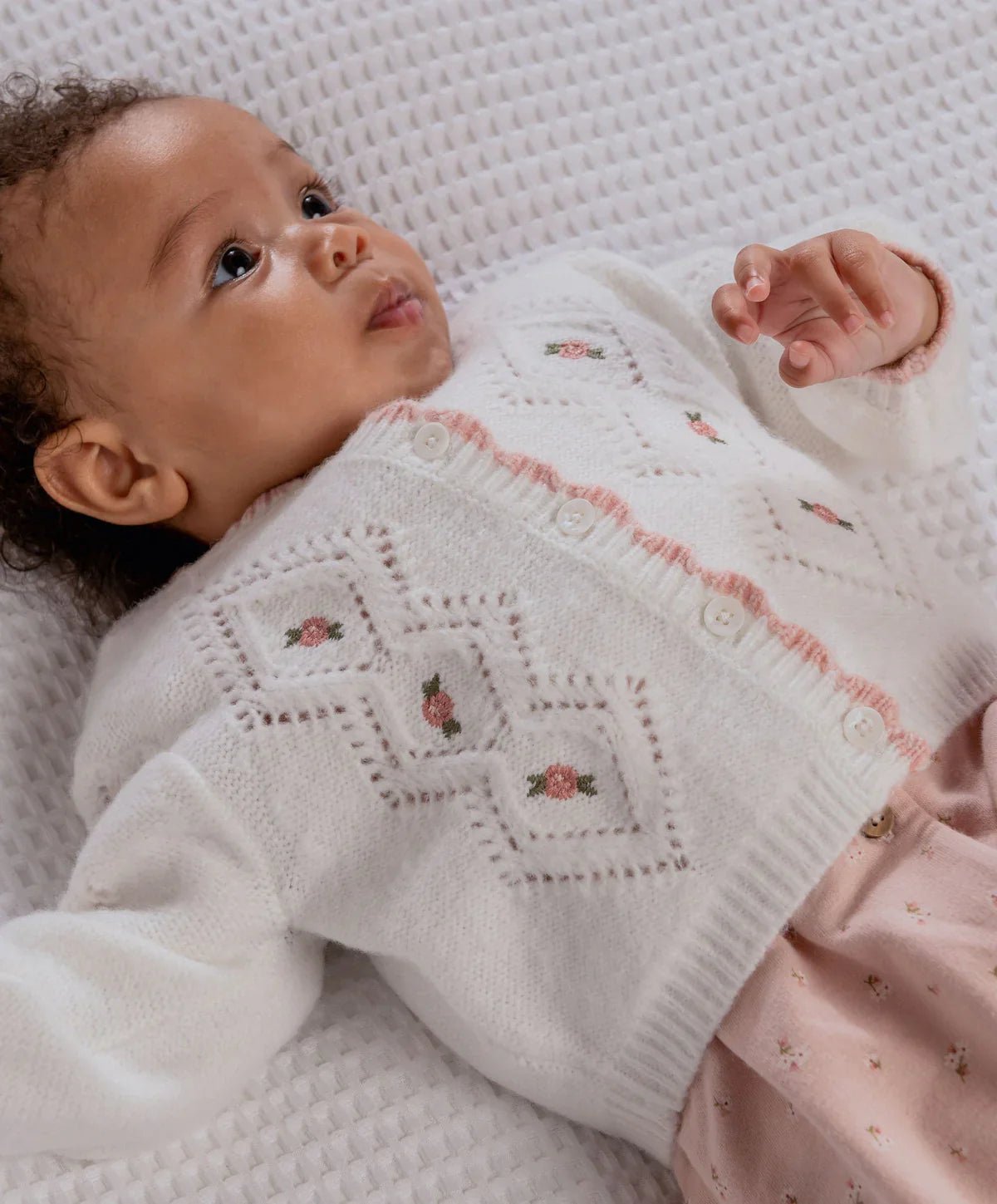 Baby wearing white embroidered cable knit cardigan with pink button placket and floral motifs