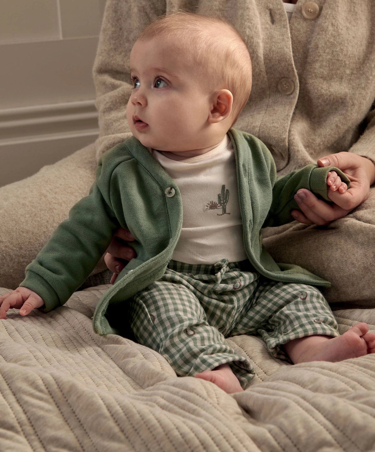Baby reclining in green button jacket with pockets, white bodysuit, and green gingham trousers.