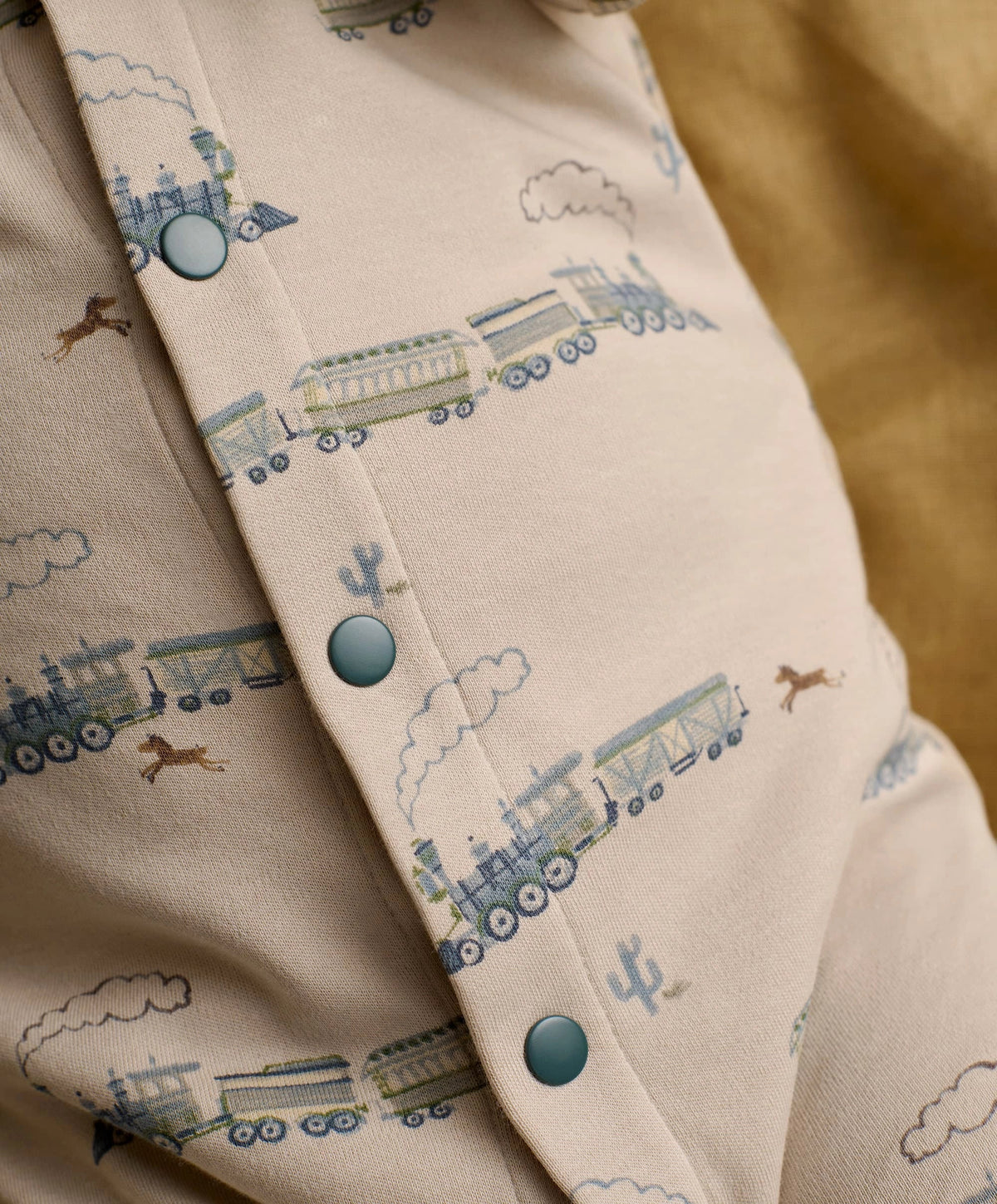 Cream long-sleeve romper with blue train print and turquoise snap buttons, close-up