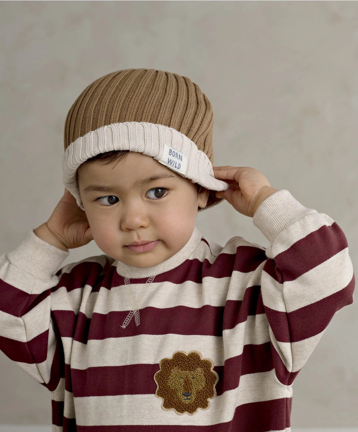 Brown knit beanie with white BORN WILD patch, worn by child holding brim