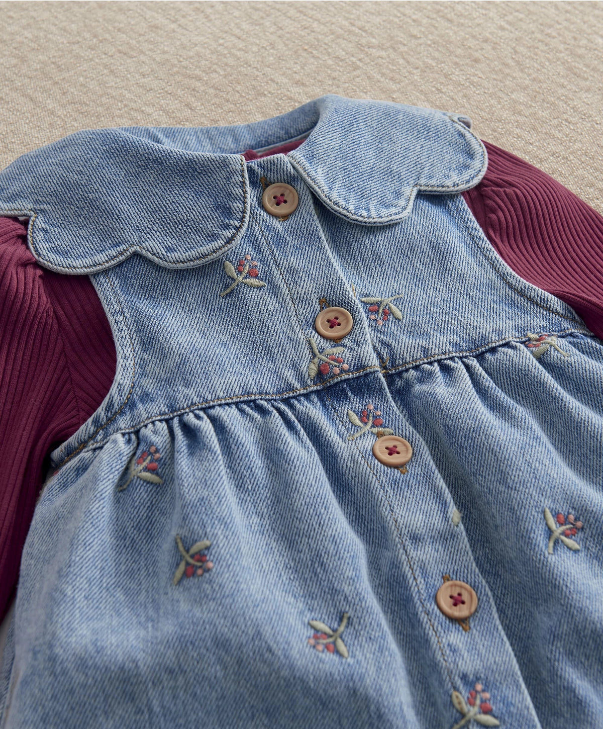 Denim dungaree bib with embroidered bows and tan buttons, burgundy ribbed sleeve cuffs