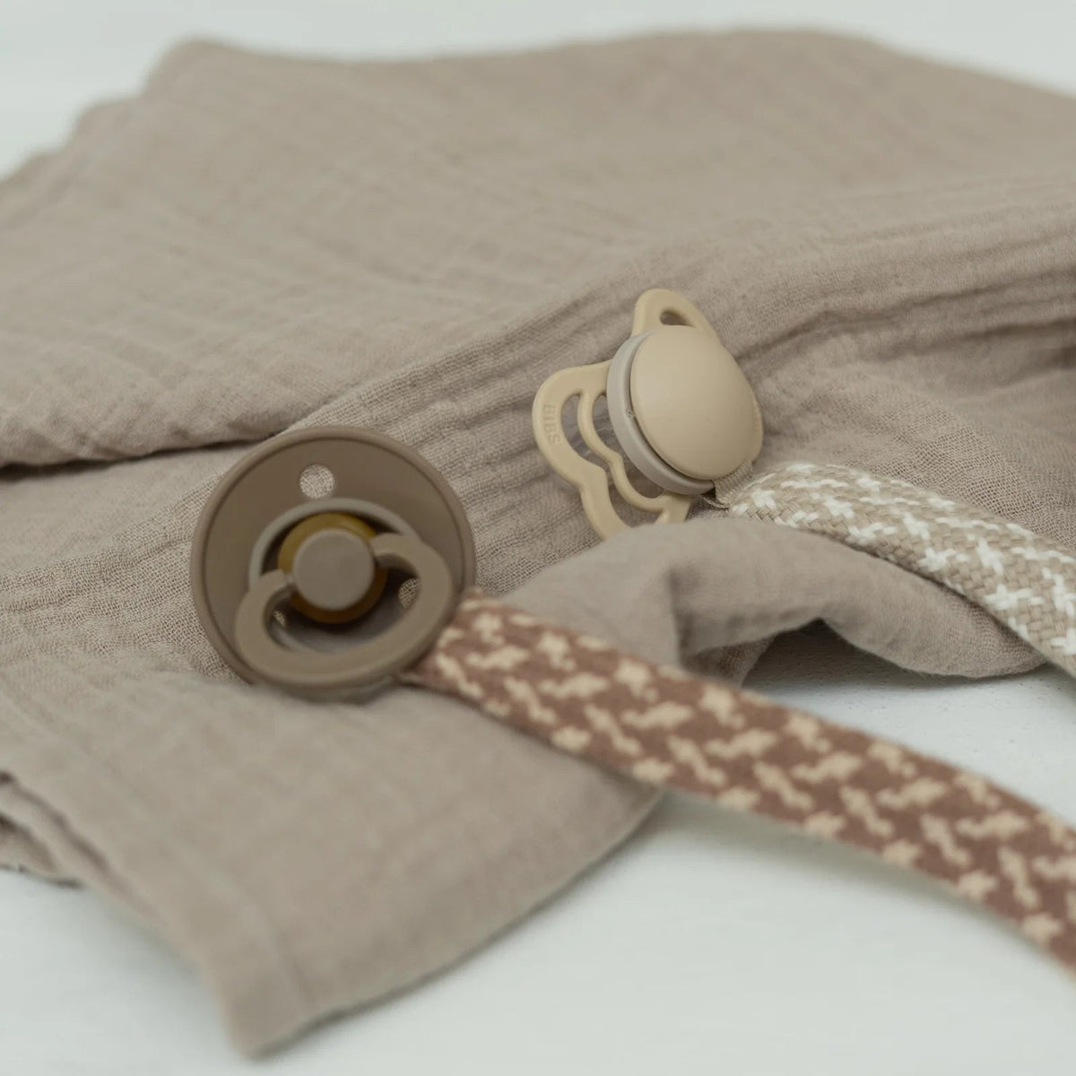 Woven pacifier clip with blush-and-ivory strap attached to two beige pacifiers