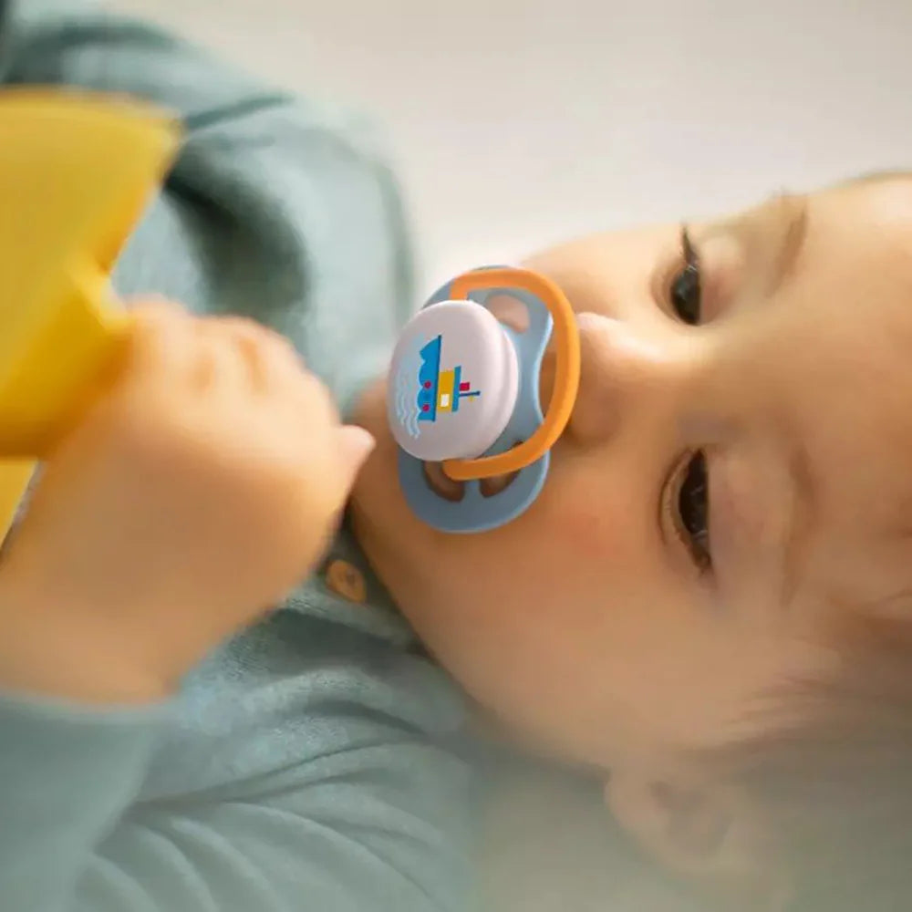 Baby lying down sucking a blue/orange pacifier with boat graphic.