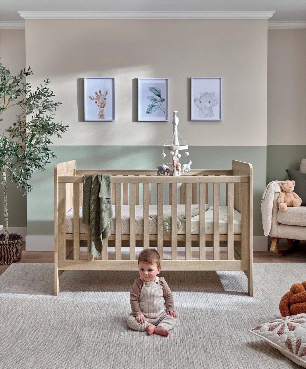 Light oak cotbed with slat rails and hanging mobile, toddler kneeling on rug