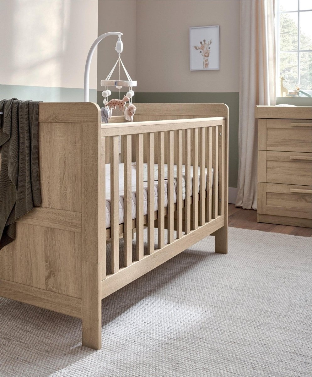 Light oak cotbed with paneled ends and vertical slat sides, plus hanging mobile