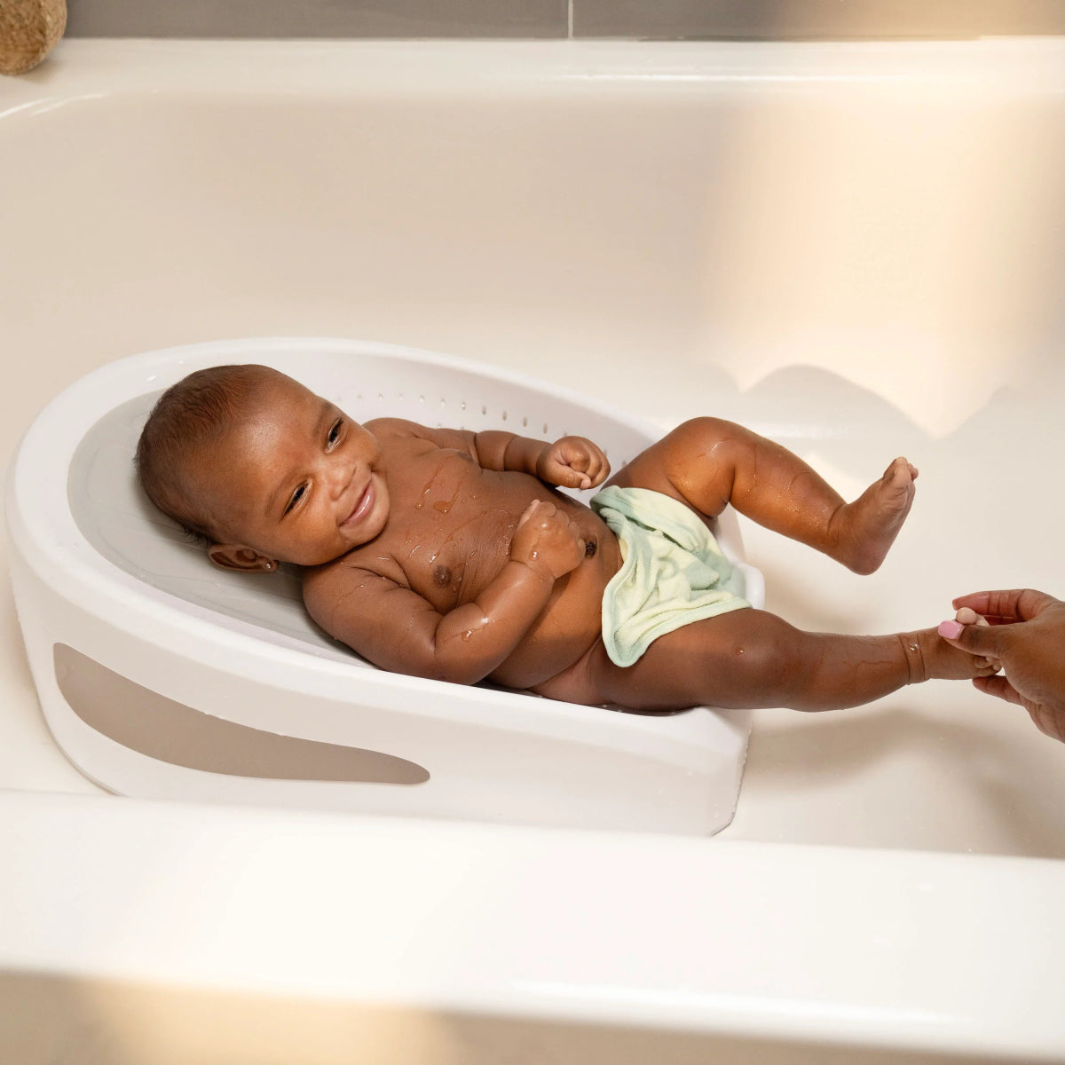 Baby lying in white bather tub with gray insert and drain holes