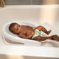 Baby reclining in white baby bather tub with gray insert and drain holes