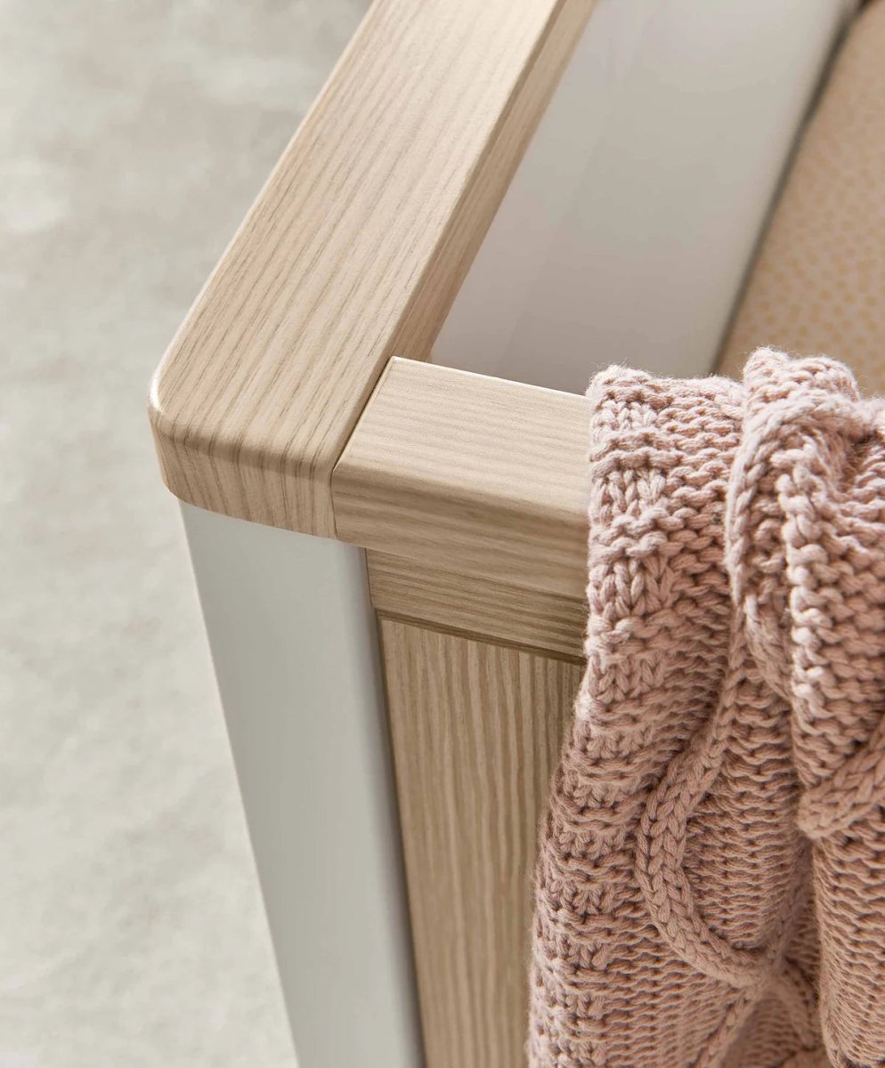 White cot side with light wood top rail, vertical slats, and knitted blanket draped