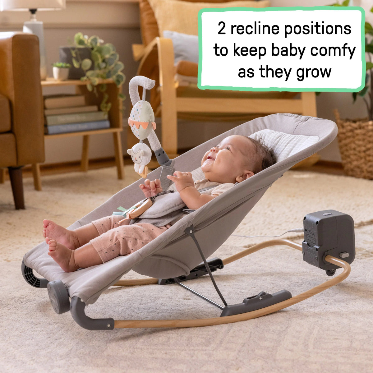 Grey baby rocking seat with baby reclined, toy overhead, and rocking base on carpet
