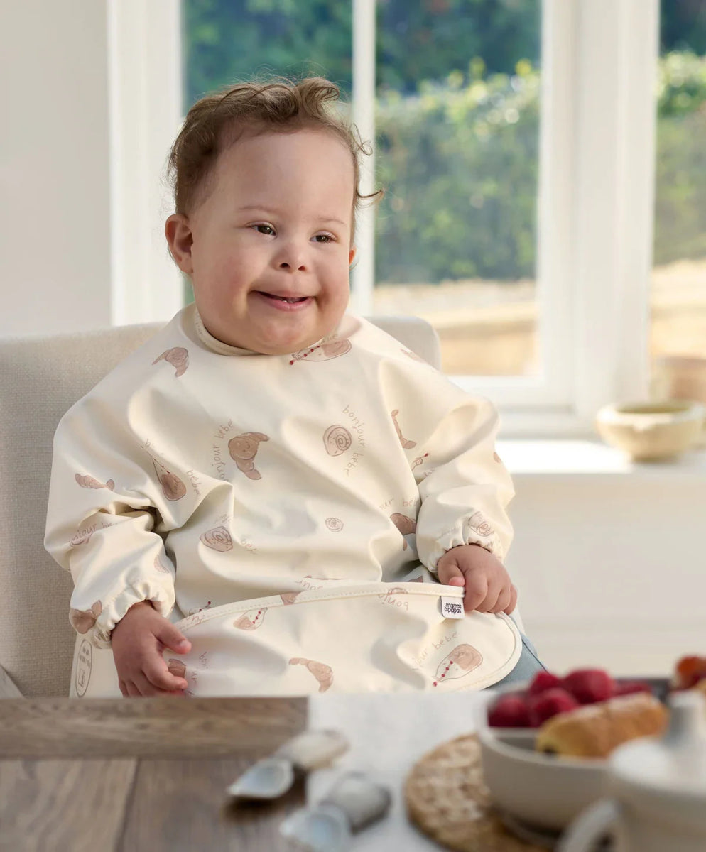 Long-sleeve baby bib in cream with animal print and bib pocket, worn by baby.