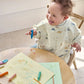Long-sleeve baby bib in cream with blue animal print, worn by a smiling child.