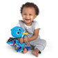 Baby in gray outfit holding blue plush sea character with striped cape and yellow bow