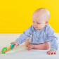 Barbell rattle toy with green end discs and orange/yellow handle, held by baby