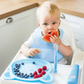 Blue suction-cup feeding spoon held near a fruit bowl