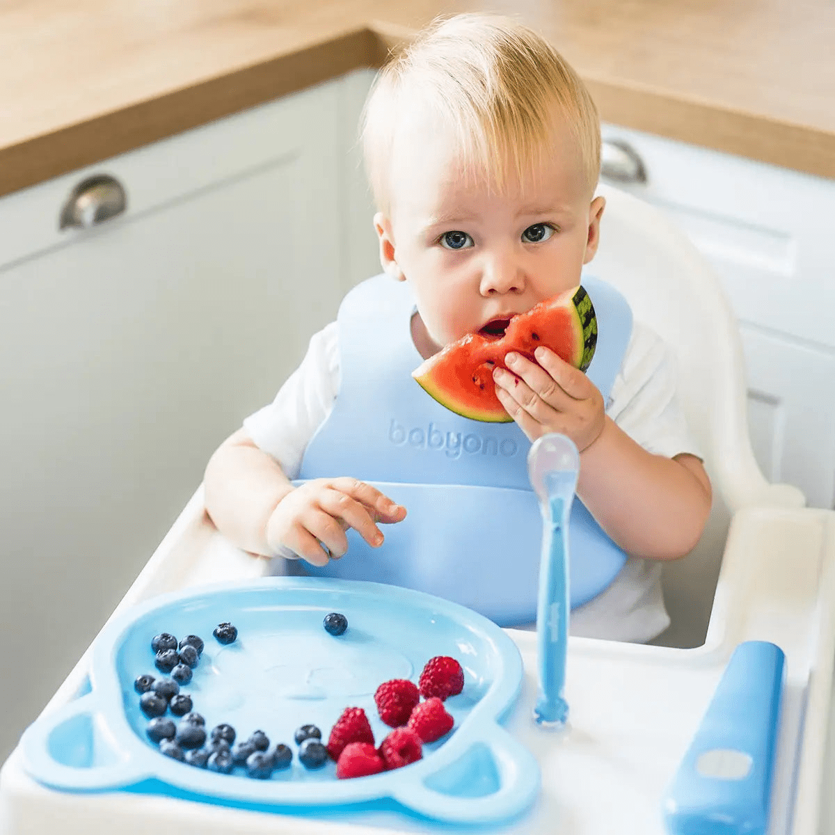 Blue suction-cup feeding spoon held near a fruit bowl