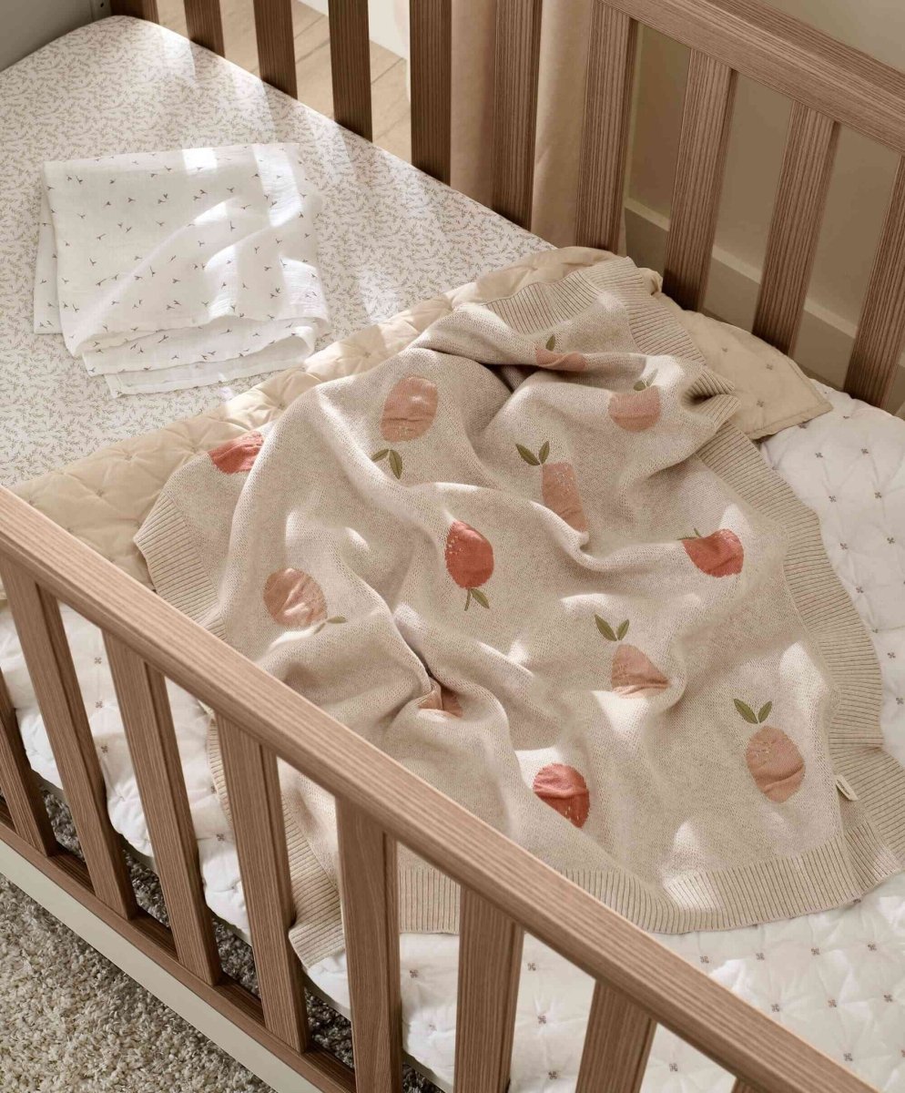 Knitted baby blanket draped over wooden crib rail, light beige with fruit and leaf pattern